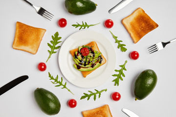 top view of toasts on plate with avocados, cherry tomatoes, arugulas leaves, forks and knives on grey background