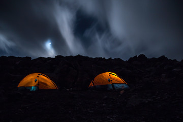 Night mountain landscape with two oranges illuminated tents. Silhouettes of mountain peaks with clouds. 