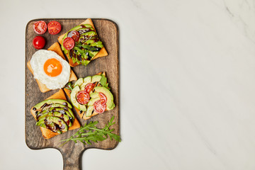 top view of wooden cutting board with toasts, scrambled egg and cherry tomatoes on marble surface