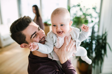 A portrait of young father with a baby girl standing indoors in a room.