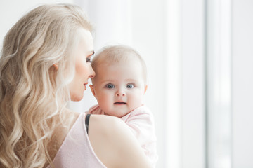 Young mother taking care of her little baby girl. Beautiful mom and her daughter indoors in the bedroom. Loving family. Attractive mum holding her child.