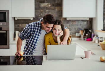 A young couple standing in a kitchen at home, using laptop.