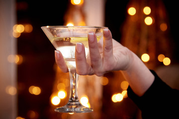 A glass with martini in a female hand on a dark background with bokeh.