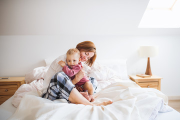 A young mother with little daughter sitting indoors on bed in the morning, playing.