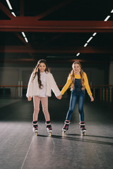 Two cute long haired children in roller skates standing in roller rink and holding hands