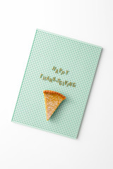 Happy thanksgiving text with a piece of pumpkin pie on a mint green pegboard on a white background, creative thanksgiving day concept, top view