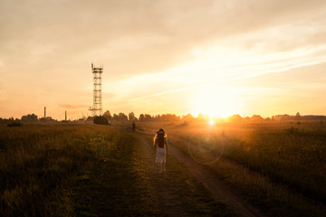 Naklejka na ściany i meble A girl with a floral wreath on her head is walking barefoot across the field at sunset.