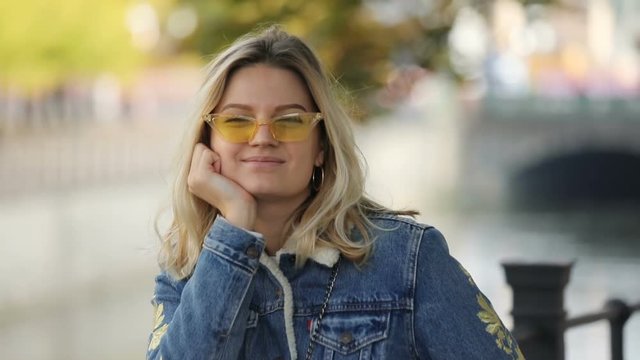 Portrait of happy pretty blonde woman wearing sunglasses smiling near a beautiful river in the city. Fall, cityscape, blurred background.