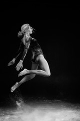Beautiful slim girl wearing a gymnastic bodysuit covered with clouds of the flying white powder jumps dancing on a dark. Artistic conceptual and advertising black and white photo.