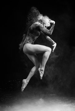 Beautiful slim girl wearing a gymnastic bodysuit covered with clouds of the flying white powder jumps dancing on a dark. Artistic conceptual and advertising black and white photo.