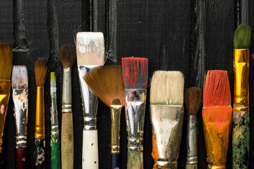 Artist's tools, brushes in paint are in a row on a black wooden background.