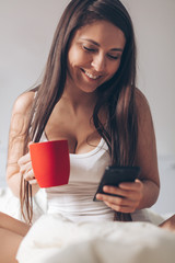 Fototapeta premium Young smiling woman drinking coffee at home in her bed and checking her smartphone