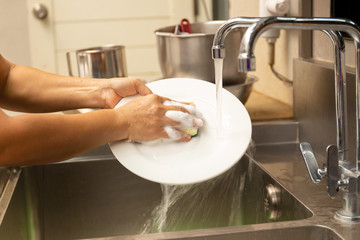 Hands washing dirty dishes with running water in kitchen sink.