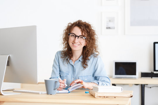 Portrait of smiling female student searches necessary information in intrenet, reads information on web page via laptop computer, writing down ideas for best education
