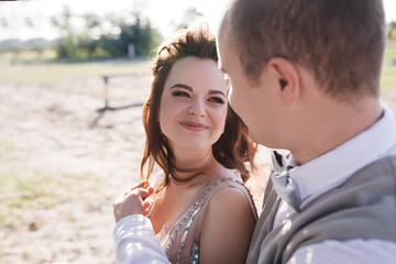 Wedding at sunset. Couple is sitting on a bench under a tree. Beige dress with sparkles. Light suit with a bow tie. The bride and groom embrace and kiss.