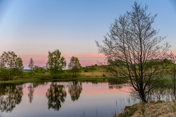 Fototapeta na wymiar A tranquil sunset scene with a pink and blue sky and trees reflected in a calm lake.