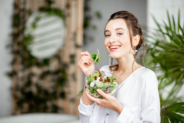 Portrait of a young woman in white shirt eating healthy salad indoors with green plants on the...