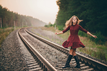 Beautiful dancing girl with curly natural hair enjoy nature in forest on railway. Dreamer lady in burgundy dress walk on railroad. Dance of inspired girl on rails at dawn. Sun in hair in autumn.