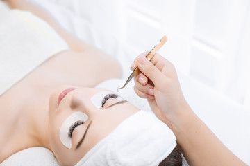 Obraz na płótnie Canvas Eyelash Extension Procedure. Close up view of beautiful Woman with Long Eyelashes. Stylist holding gold tweezers, tongs and making lengthening lashes for girl in a beauty salon. Beauty Concept.
