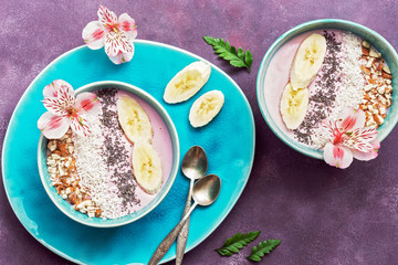 Fresh healthy breakfast - yogurt with banana, almond,coconut flakes, chia seeds in a blue bowl decorated with flowers on a purple background. Top view