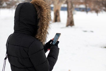 Upper body shot of woman with fur hooded jacket holding smart phone in her hand. Cold weather with snow all around. Turned back to camera.