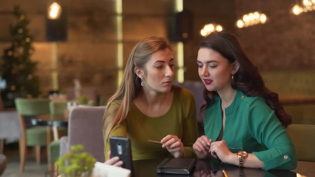 Two charming young women are sitting in a cafe indoors and talking in a video chat with someone. Both women are doing some business and looking totally serious.