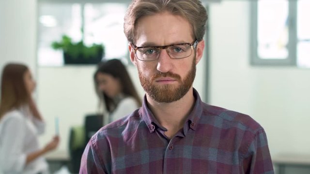 Portrait of a bearded guy in glasses and in a casual plaid shirt standing in the office center in the lobby on the background of two girls office workers. The man smiles first and then becomes serious