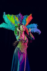 Beautiful young woman in carnival peacock costume. Beauty model woman at party over holiday background with magic glow. Christmas and New Year celebration. Glamour lady with perfect make up and