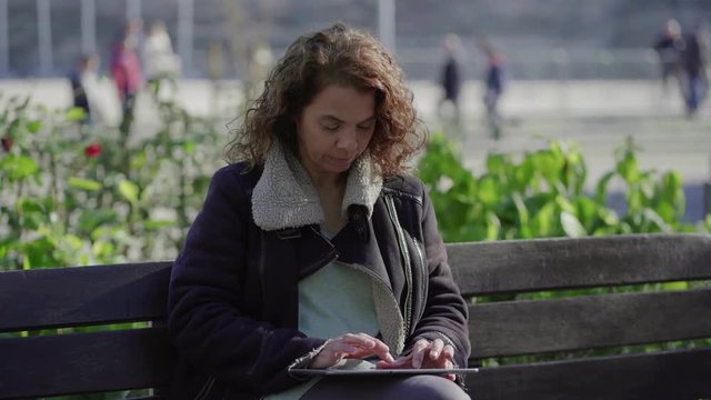 Focused mature woman using tablet outdoor. Curly midle aged lady wearing warm jacket sitting on wooden bench in park and typing on tablet. Technology concept