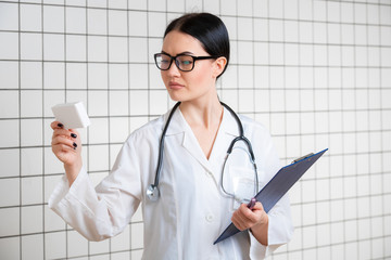 A friendly cute girl with dark hair and glasses, wearing a lab coat, reads carefully the information on the pack in a modern pharmacy.