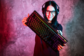 Nerd geek young adult women holding gaming keyboard over colorful pink and blue neon lit wall. Gaming gamers concept.