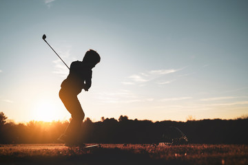 Young junior golfer practicing in a driving range with beautiful sunset light in winter. - obrazy, fototapety, plakaty