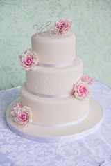 Fototapeta na wymiar White three-tiered wedding cake with pink roses made of mastic stands on the table a delicate green background. letters bride and groom