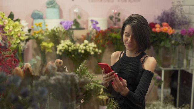 Beautiful slim thin hot sexy brunette in black shoulderless sweater touching different flowers in flower shop while doing videocall. Girl being mesmerized with flowers