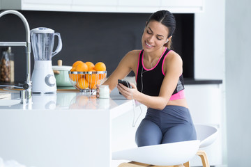 Sporty young woman listening to music with mobile phone while eating yogurt in the kitchen at home.