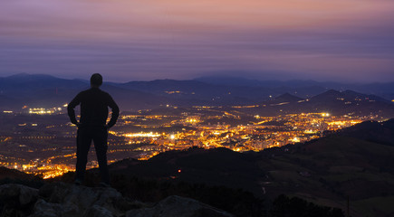 A man stands on top of a mountain, against the backdrop of the lights of the night city.