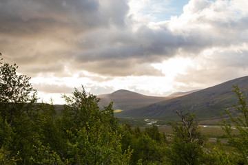 Panorama view of mountains behind Kebnekaise mountain station at the Kungsleden (Kings trail) trail in northern Sweden. Bright sky with clouds and green trees. 