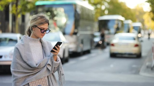 Portrait of young attractive blonde girl wearing coat and sunglasses staring at her smartphone on street. Pretty smiling woman with mobile phone standing on the city road. Traffic in the background.