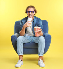 Man with 3D glasses, popcorn and beverage sitting in armchair during cinema show on color background © New Africa