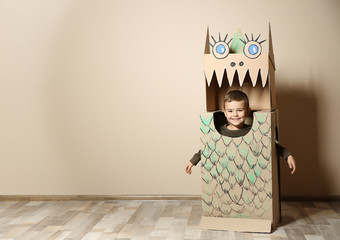 Cute little boy in cardboard costume of dinosaur near color wall. Space for text