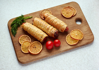 Baked sausages wrapped in puff pastry on a wooden cutting board accompanied by parsley,tomatoes and dried orange slices.Pigs in blanket,sausage rolls/Fried dough enclosed sausages