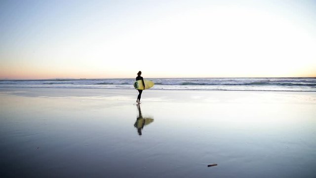 Young girl holding surfboard on the beach. Woman walking with surf into the ocean. Beautiful sunset, wind is blowing.
