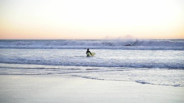 Young girl holding surfboard on the beach. Woman walking with surf into the ocean. Beautiful sunset, wind is blowing.