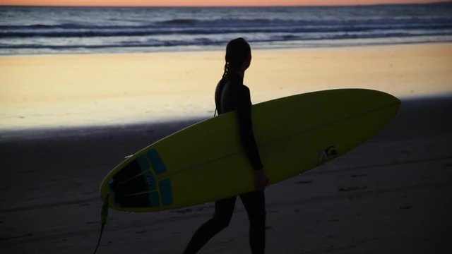 Young girl holding surfboard on the beach. Woman walking with surf along the coast. Silhouette of the woman. Beautiful sunset, wind is blowing. Relax after serfing.