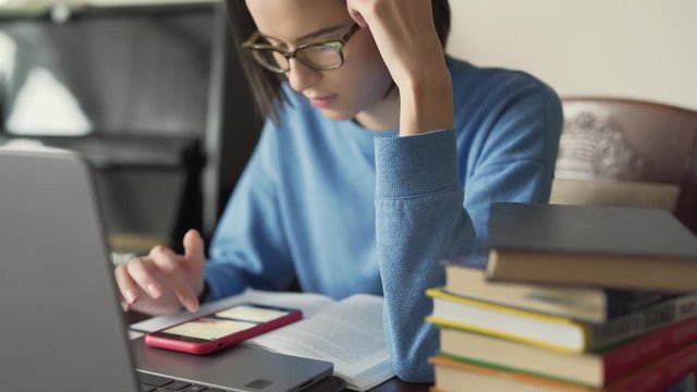 Procrastination. Cute girl with glasses is sitting in a room and watching an Instagram stories. Girl is typing on her phone and has a silver laptop and pile of books in front of her.