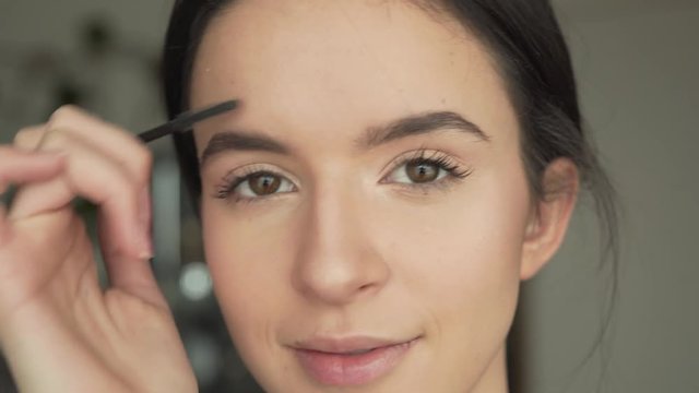 Close-up portrait of a young beautiful girl's face, who is combing her eyebrows with a brush, looking directly at the camera and doing a make-up.