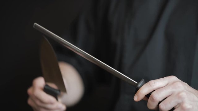 Close-up of a professional chef's hands using a honing steel rod to sharpen a knife blade on a black background