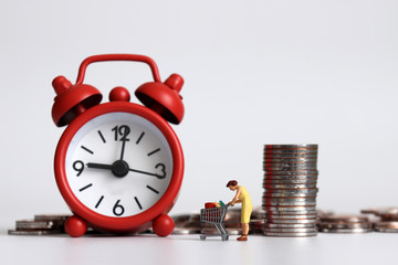 Miniature woman pulling shopping cart with a pile of coins in front of red alarm clock.