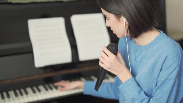 Charming girl is sitting in the room, wearing earphones, playing the piano and singing in a microphone. Girl is looking relaxed and addicted to her singing.