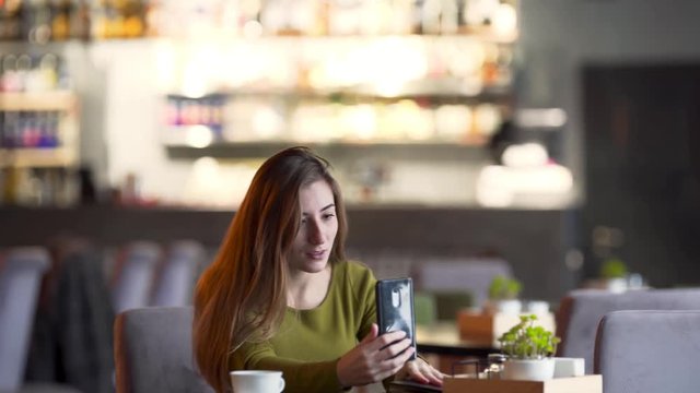Young adorable woman is sitting in a cafe and talking in a video chat with her friend. She is looking absolutely relaxed and easy.
